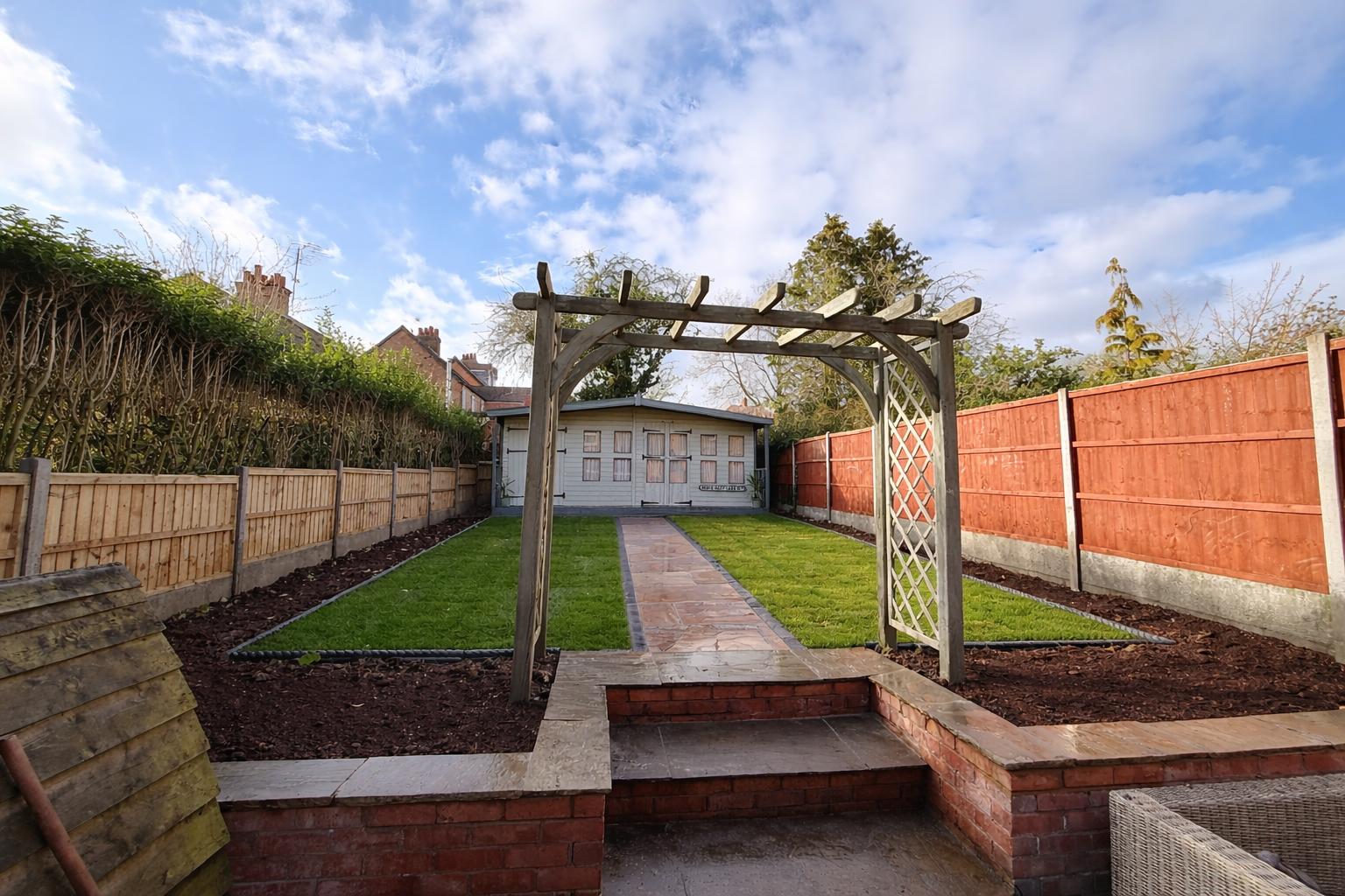Wide view of a landscaped residential garden featuring a wooden pergola, brick steps, sandstone pathway, and fresh turf in Market Harborough by AAS Group Construction
